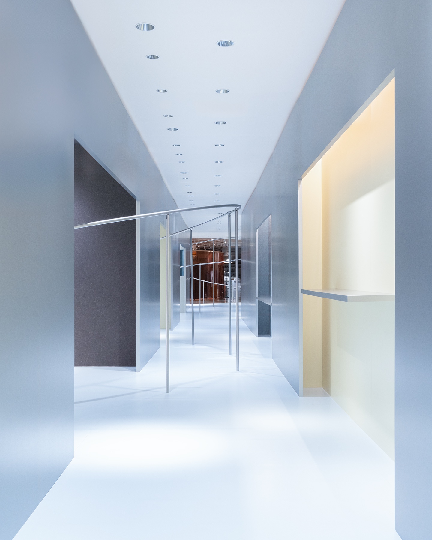 A modern, minimalist corridor with light gray walls, a white floor, and a curving silver metal rail. Recessed lights line the ceiling, with warm-toned accents on walls.