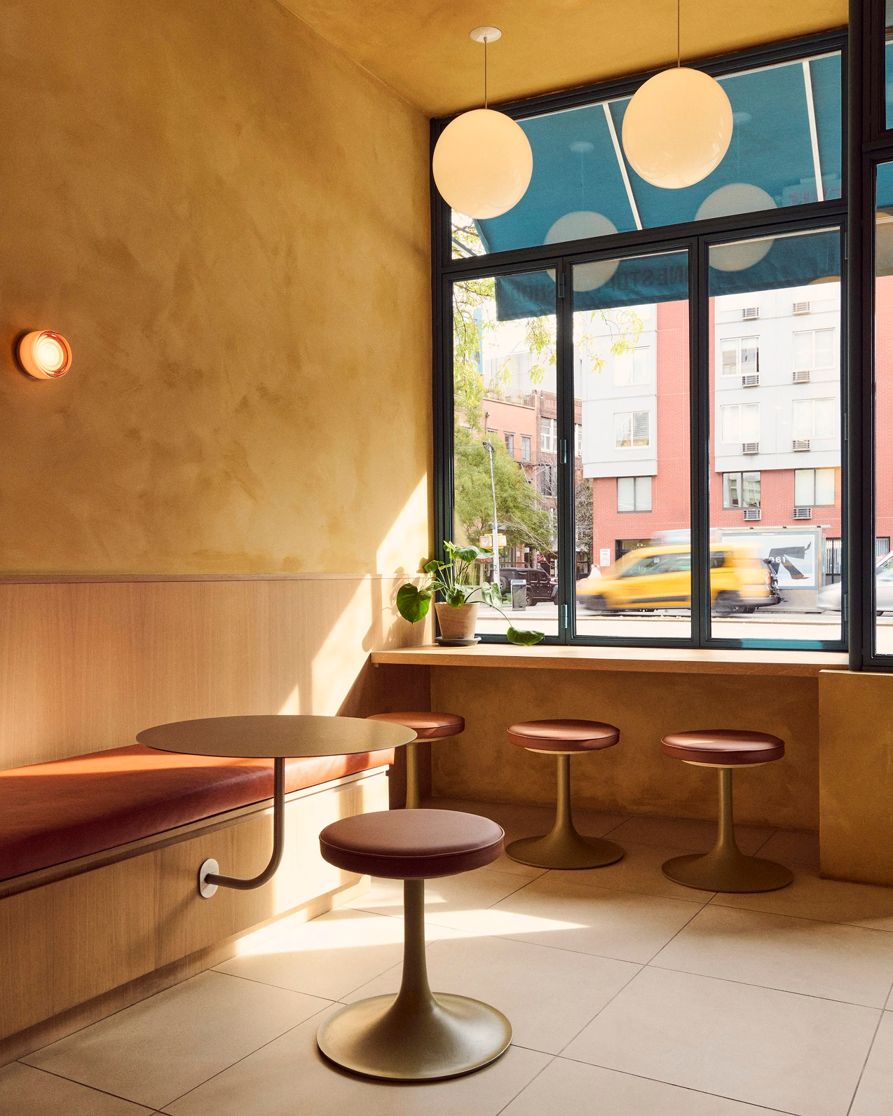Modern cafe interior with ochre walls, wooden bench seating, round table, and stools next to large street-view windows.