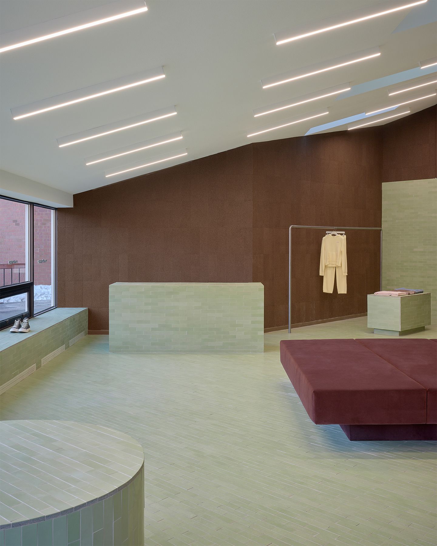 A modern retail space with brown walls, light green tiled floors, a long green counter, a maroon bench, and linear ceiling lights.