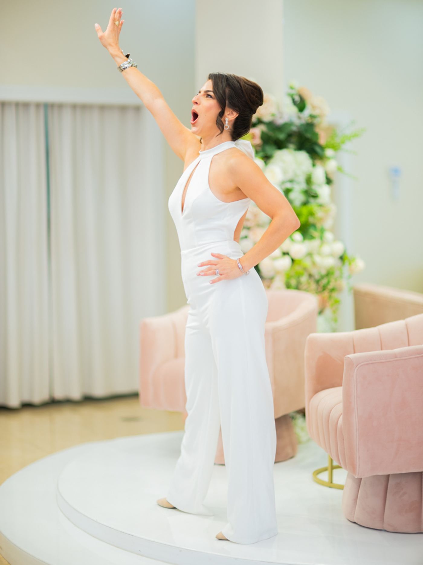 Renée Marino in a white jumpsuit, arm thrown skyward mid-exclamation, energizing the room