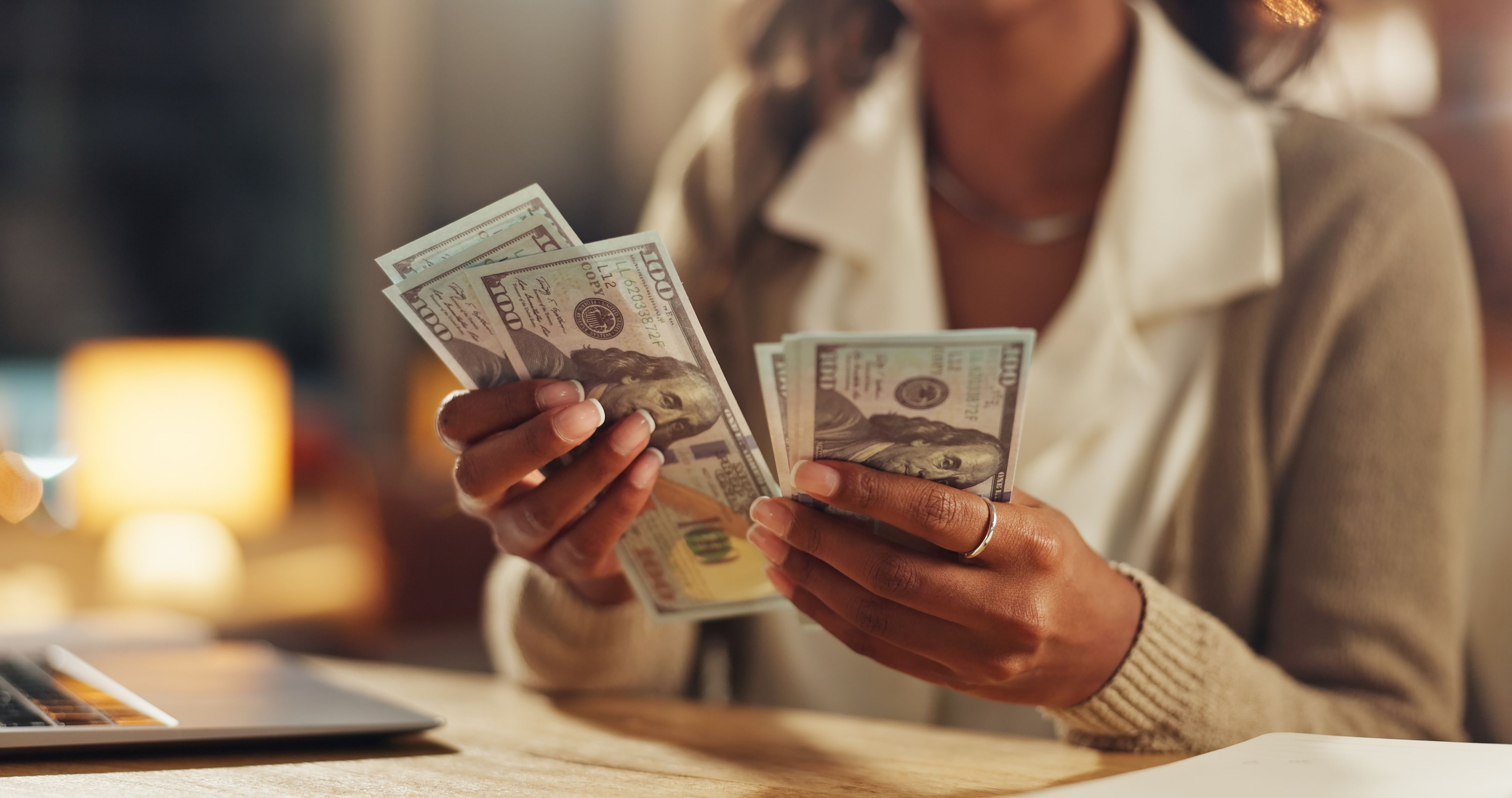 Woman counting U.S. dollar bills in her hands, including hundred dollar bills, at a desk with warm lighting in the background – representing budgeting for immigration application costs.