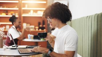 J2 Visa Holder Working Remotely With Ead Work Permit | Immiva Young professional with curly hair working on laptop at a coffee shop, representing J2 visa holders who can work freely in the U.S. after obtaining their EAD work permit