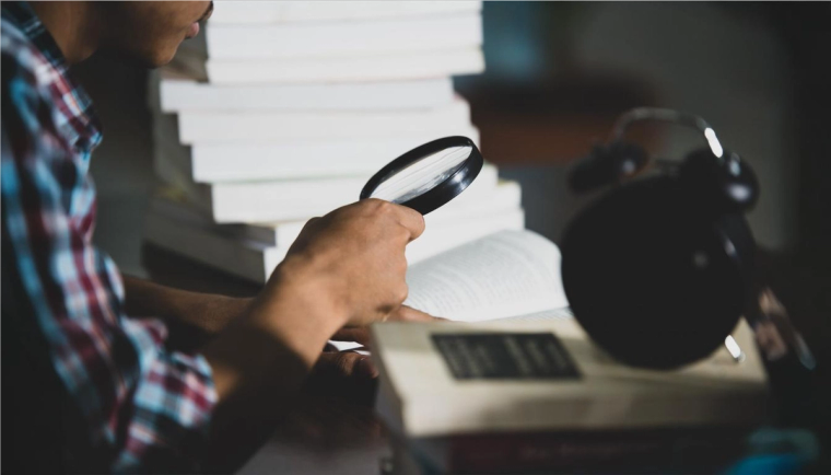 Searching the Green Card Number with Magnifying Glass and Book | Immiva Person examining a green card with a magnifying glass surrounded by stacked books and a clock on the desk.