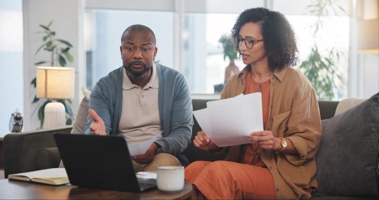 Couple reviewing I-751 documents to remove conditions on green card | Immiva A couple sits together on a couch reviewing Form I-751 paperwork and using a laptop to file their petition to remove conditions on their marriage-based green card