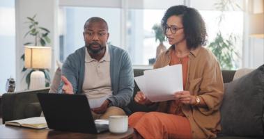 A couple sits together on a couch reviewing Form I-751 paperwork and using a laptop to file their petition to remove conditions on their marriage-based green card