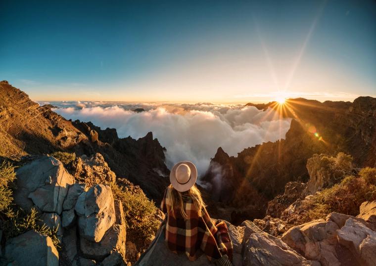 Woman Watching Sunrise Over Mountains While I-751 Is Pending | Immiva Woman wearing a blanket and hat watching the sunrise over clouds from a mountaintop, illustrating travel risks while an I-751 petition is pending