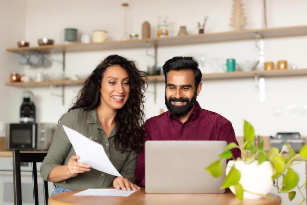 Smiling couple at a kitchen table reviewing Form I-539 on a laptop, holding documents while preparing an H-4 visa change of status application.
