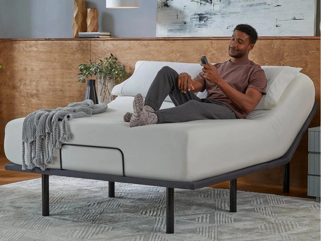 Man lounging on a bed with the head raised by the Silver base, holding the remote