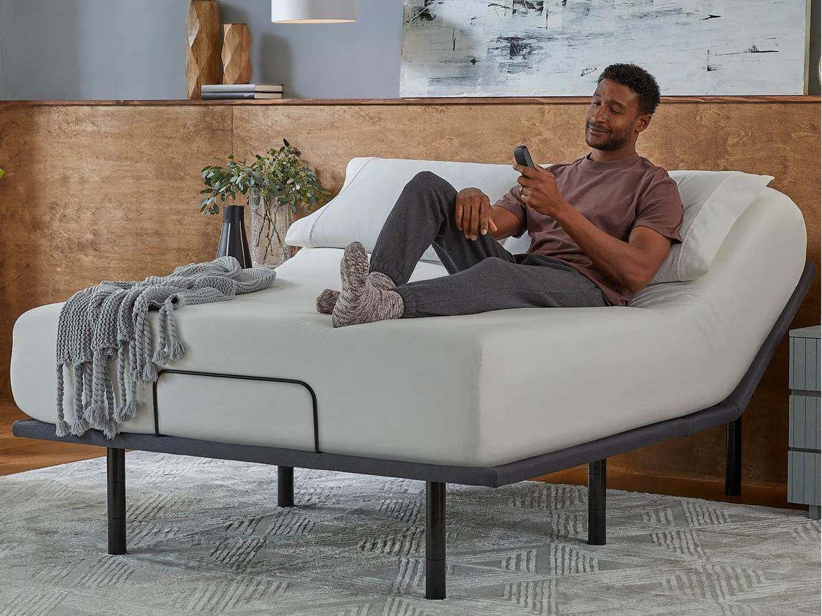 Man lounging on a bed with the head raised by the Silver base, holding the remote