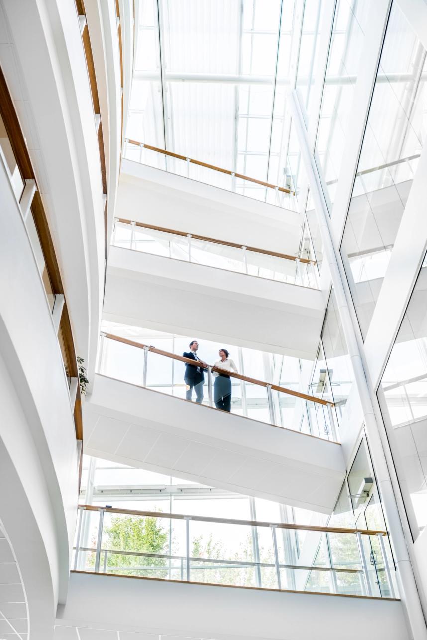 A photo of a bright, white hallway. The camera is facing up towards a couple talking on a stairway a few levels up.