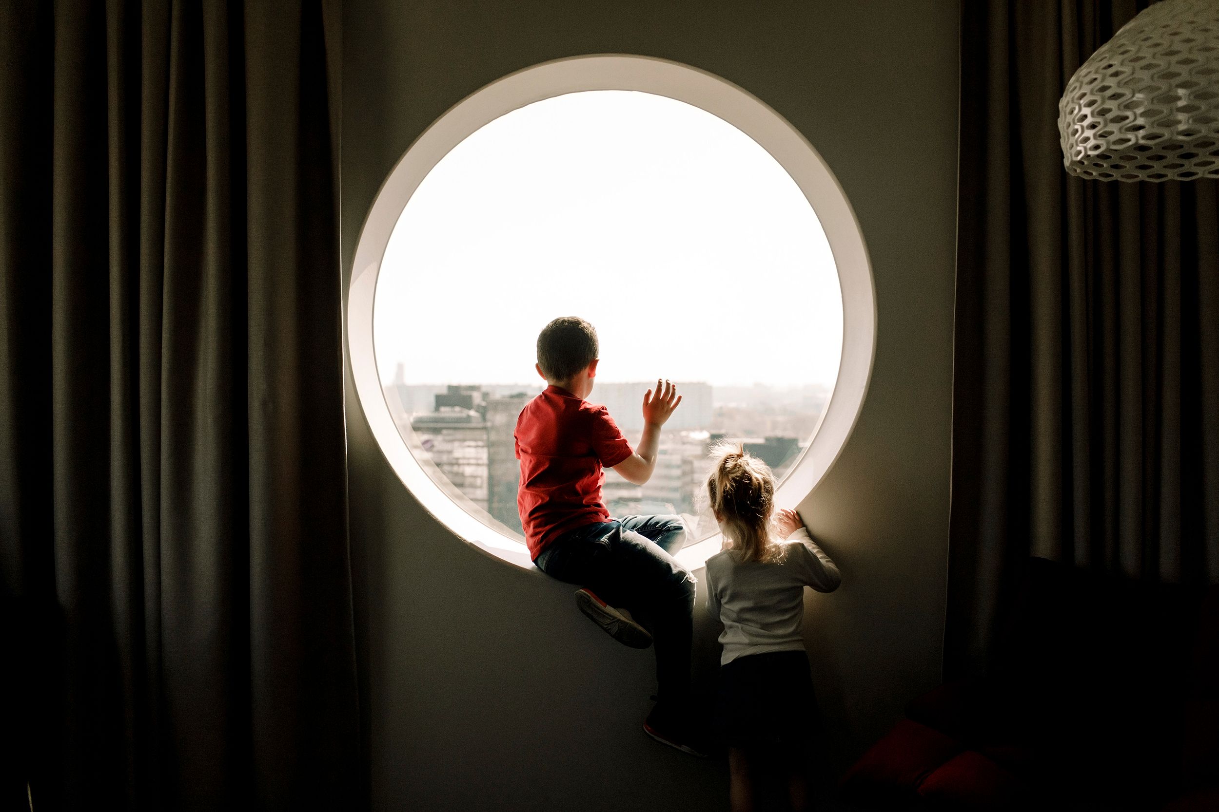 A wide, round window. A person is sitting backlit in the window sill, looking out. A child is standing on the side, also looking out.