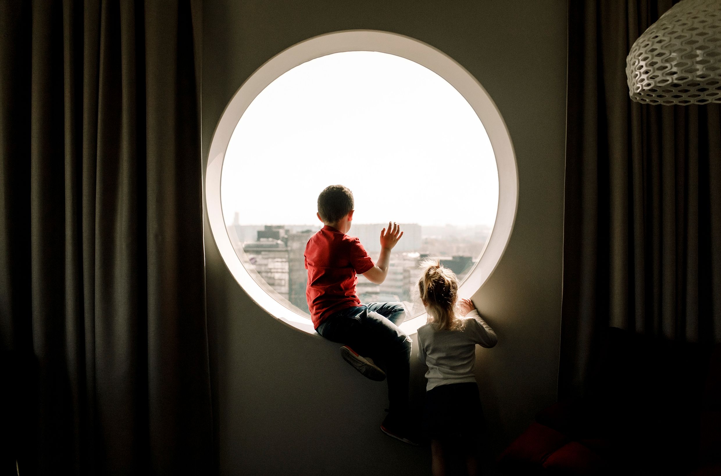 A wide, round window. A person is sitting backlit in the window sill, looking out. A child is standing on the side, also looking out.