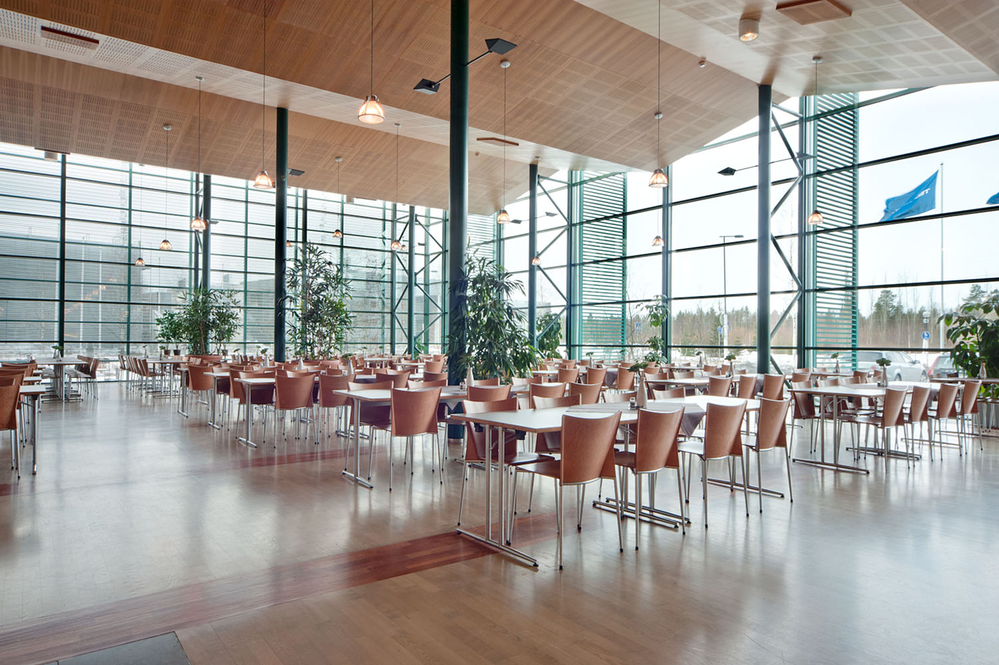 A grand, open room with tables and chairs. The walls are made of glass, the ceiling is high and vaulted. The chairs and tables are a soft brown.