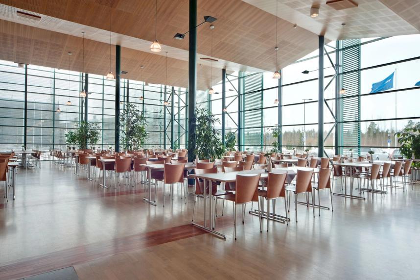 A grand, open room with tables and chairs. The walls are made of glass, the ceiling is high and vaulted. The chairs and tables are a soft brown.