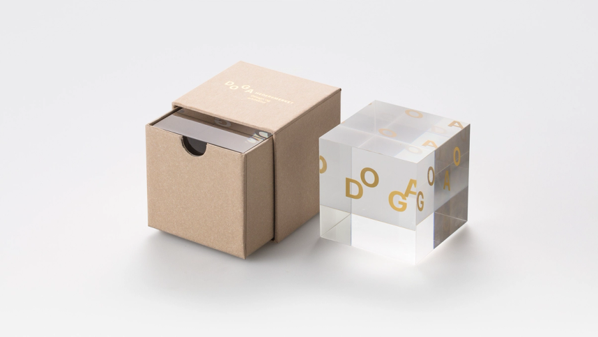 An example of DOGA awards packaging. A square cardboard box with the DOGA interface, and a glass cube award with DOGA written in gold.