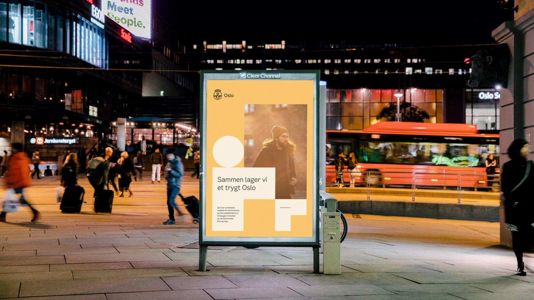 An advertisement board in a busy street. The board shows the Oslo Municipality design formula in yellow, with L-shapes and circles.