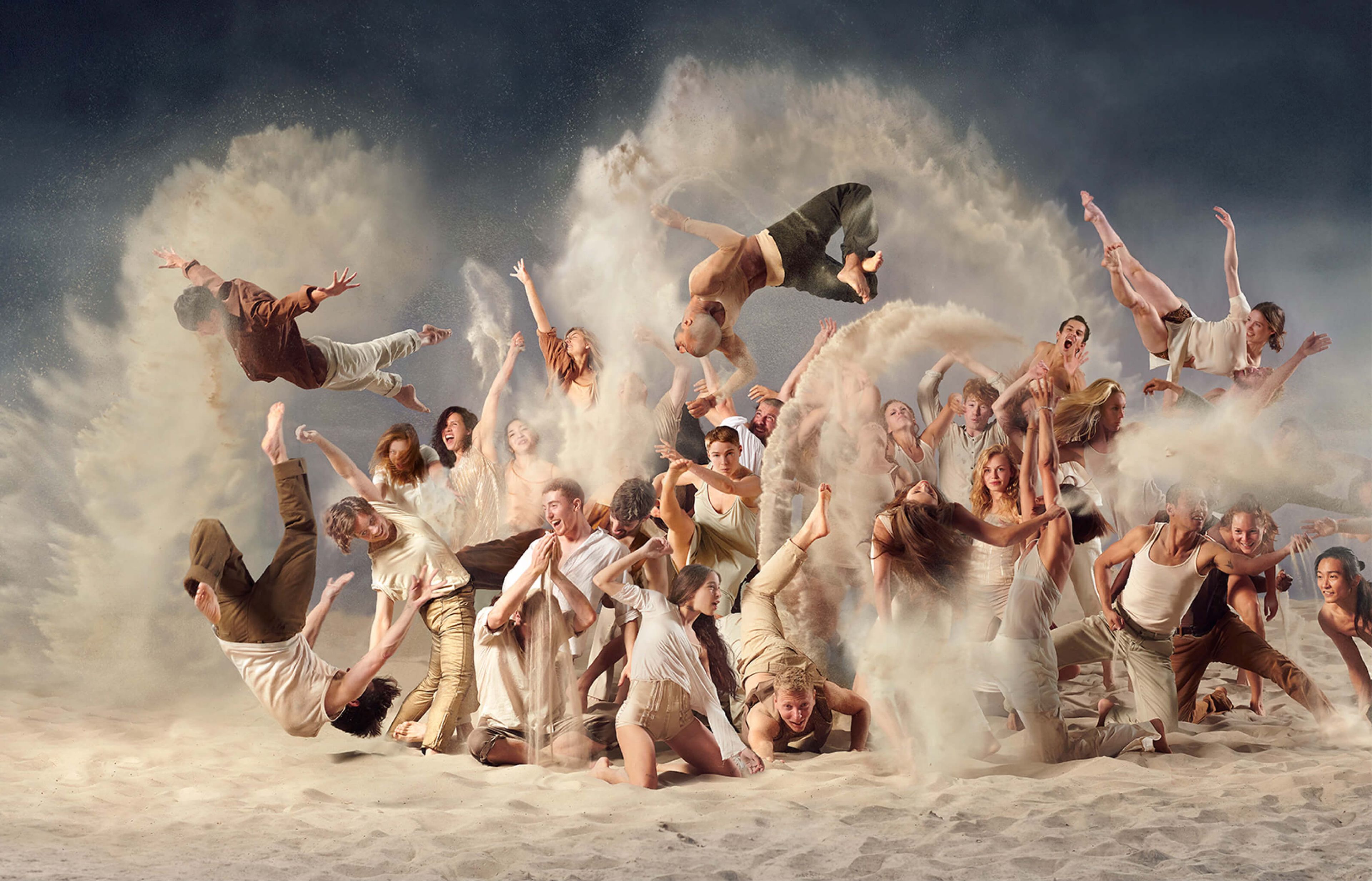 An illustrative photo from a show production, showing a crowd of people dressed in whites and browns, playing and dancing in sand