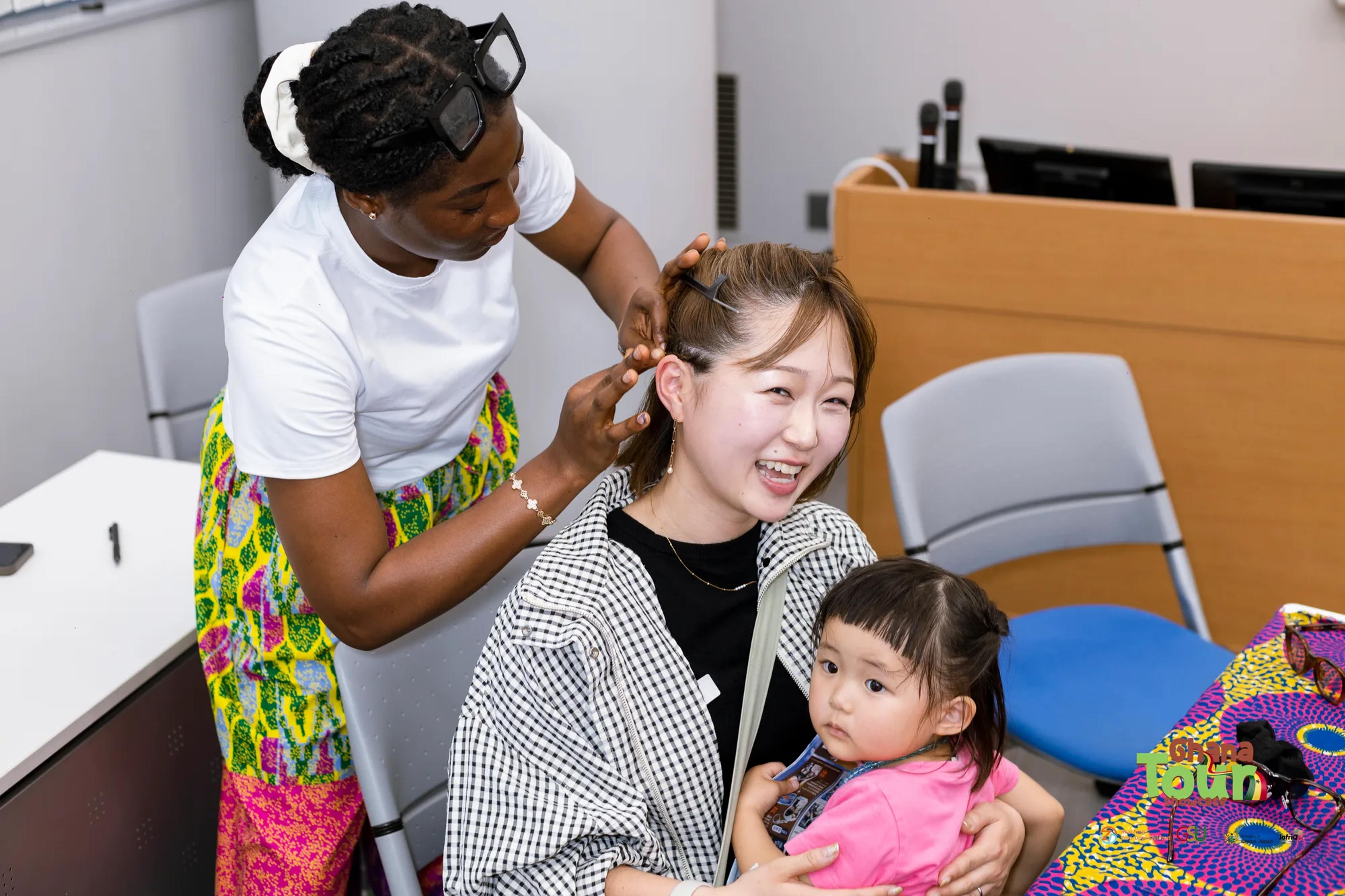 Smiling visitor holding her baby has her hair braided by a Ghanaian volunteer