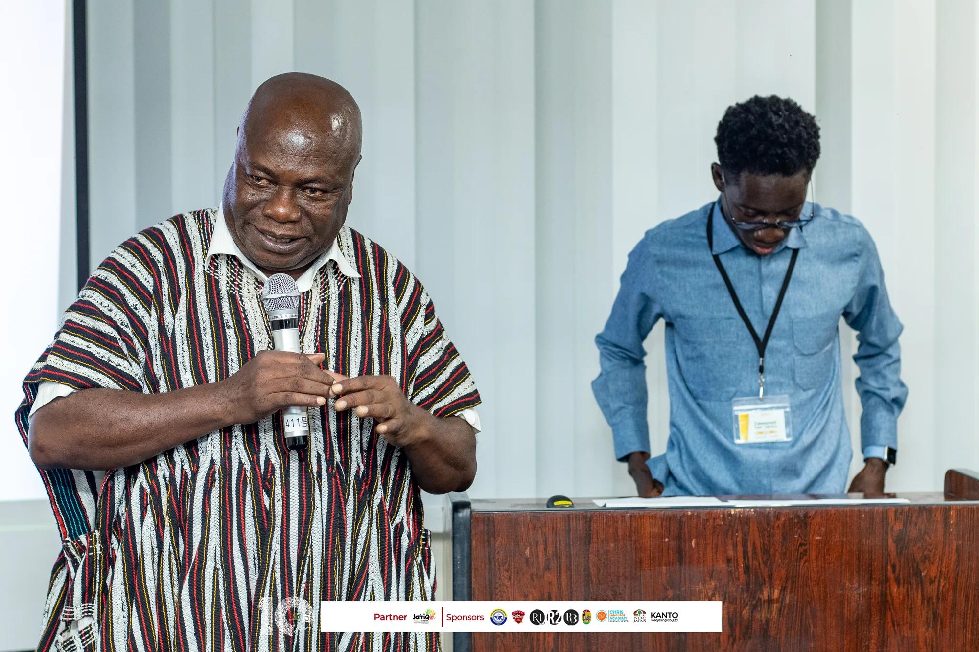 Speaker in a smock talks while a presenter prepares at the podium