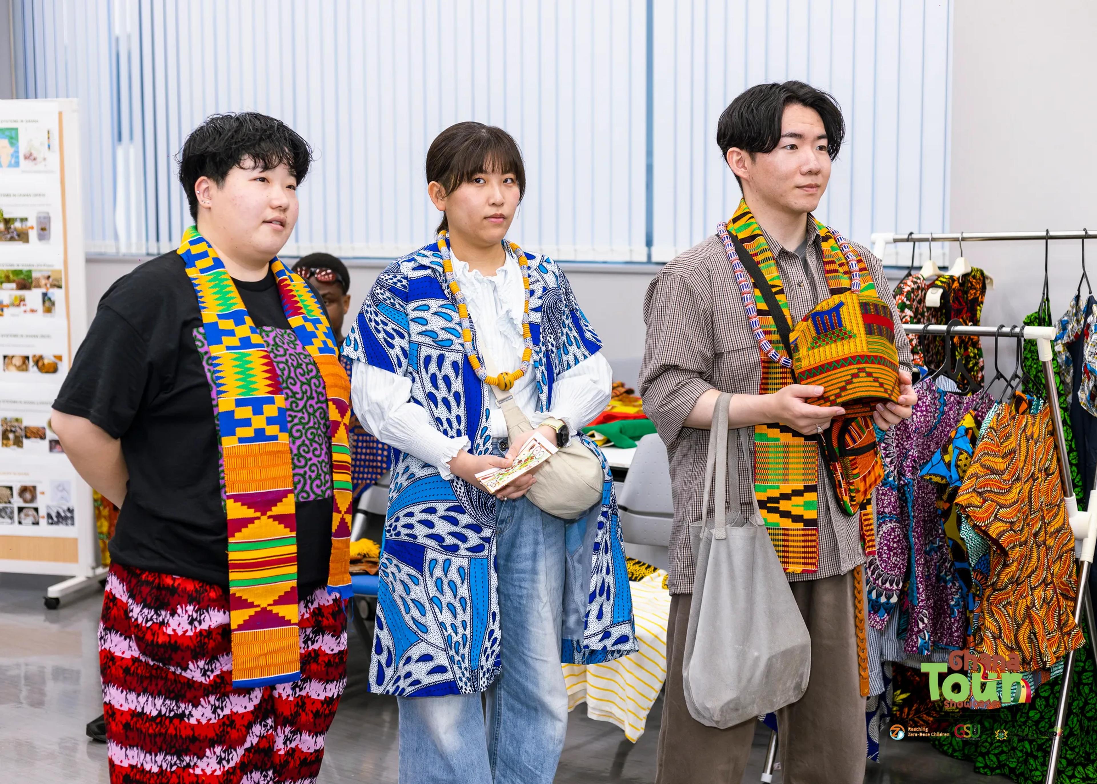 Three visitors wear kente stoles and print scarves beside the fashion rack
