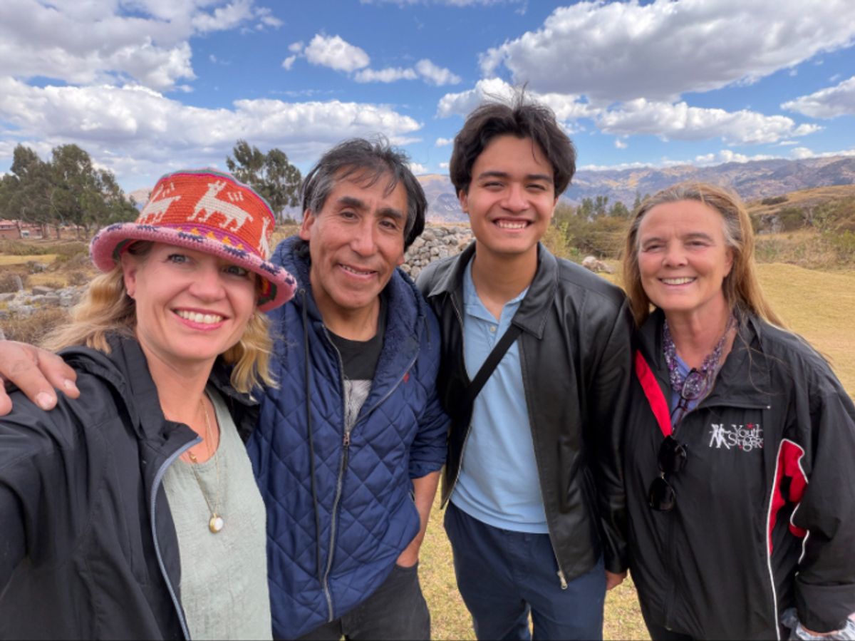 Four people pose outdoors on a sunny day, smiling at the camera on a grassy hillside with mountains and a blue sky in the background; one wears a red hat and another wears a dark jacket.