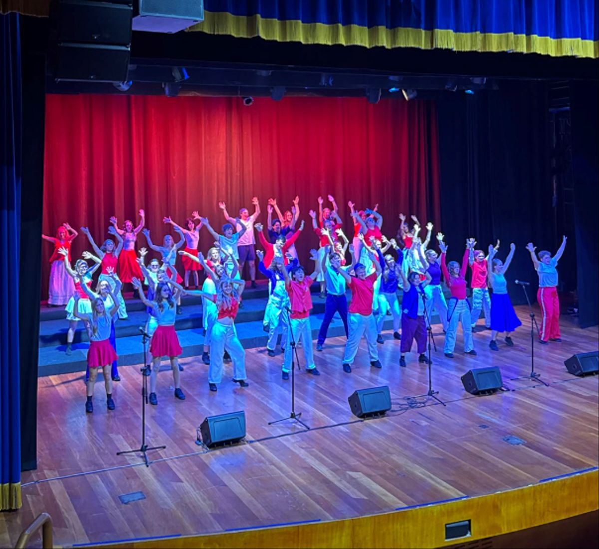A large group of performers in colorful costumes dance on a stage under red and blue spotlights, arms raised in synchronized poses with microphones and speakers along the front.
