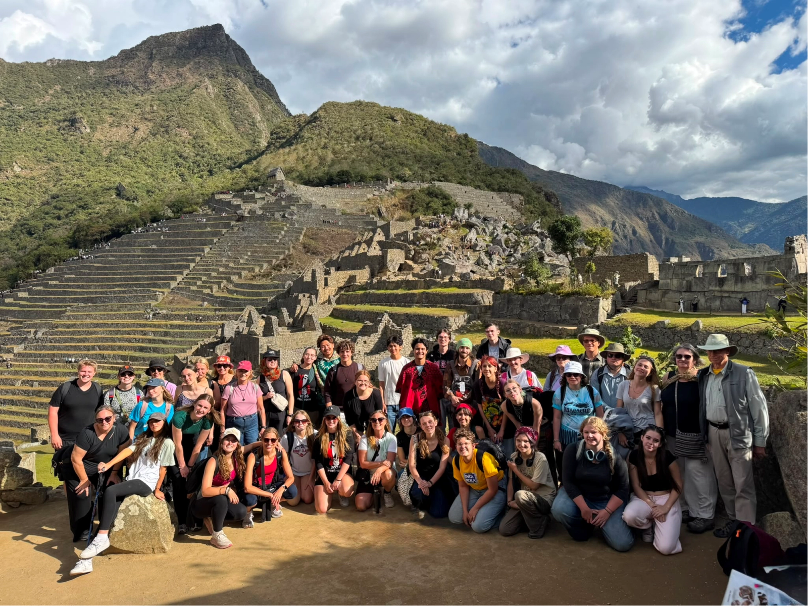 Group photo of travelers posing on a dirt path with grassy mountains and terraced ruins behind them under a partly cloudy blue sky.