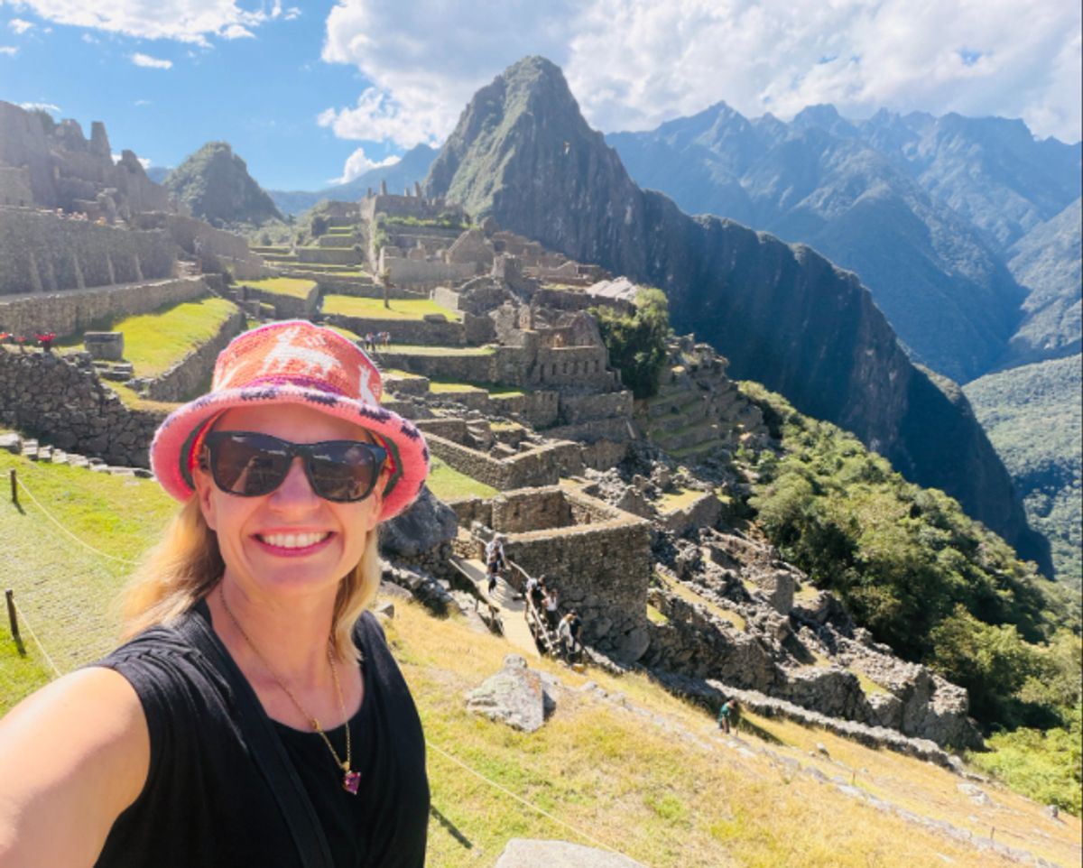A smiling person in a red sun hat and sunglasses stands in the foreground while ancient stone terraces and ruins stretch along a mountain valley under bright blue skies.