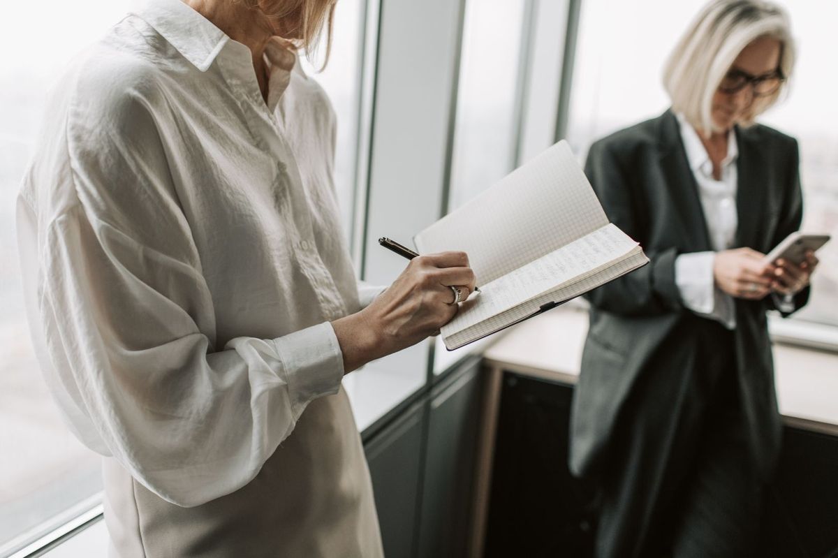 One person in a white shirt writes in a notebook while another person in a black suit looks at their phone.