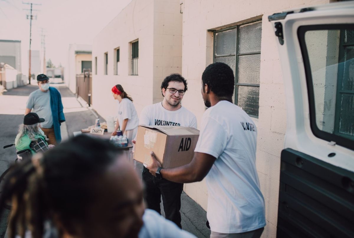 People in a sunny alleyway loading cardboard boxes from a white van, with one person holding a box while others stand nearby and talk.