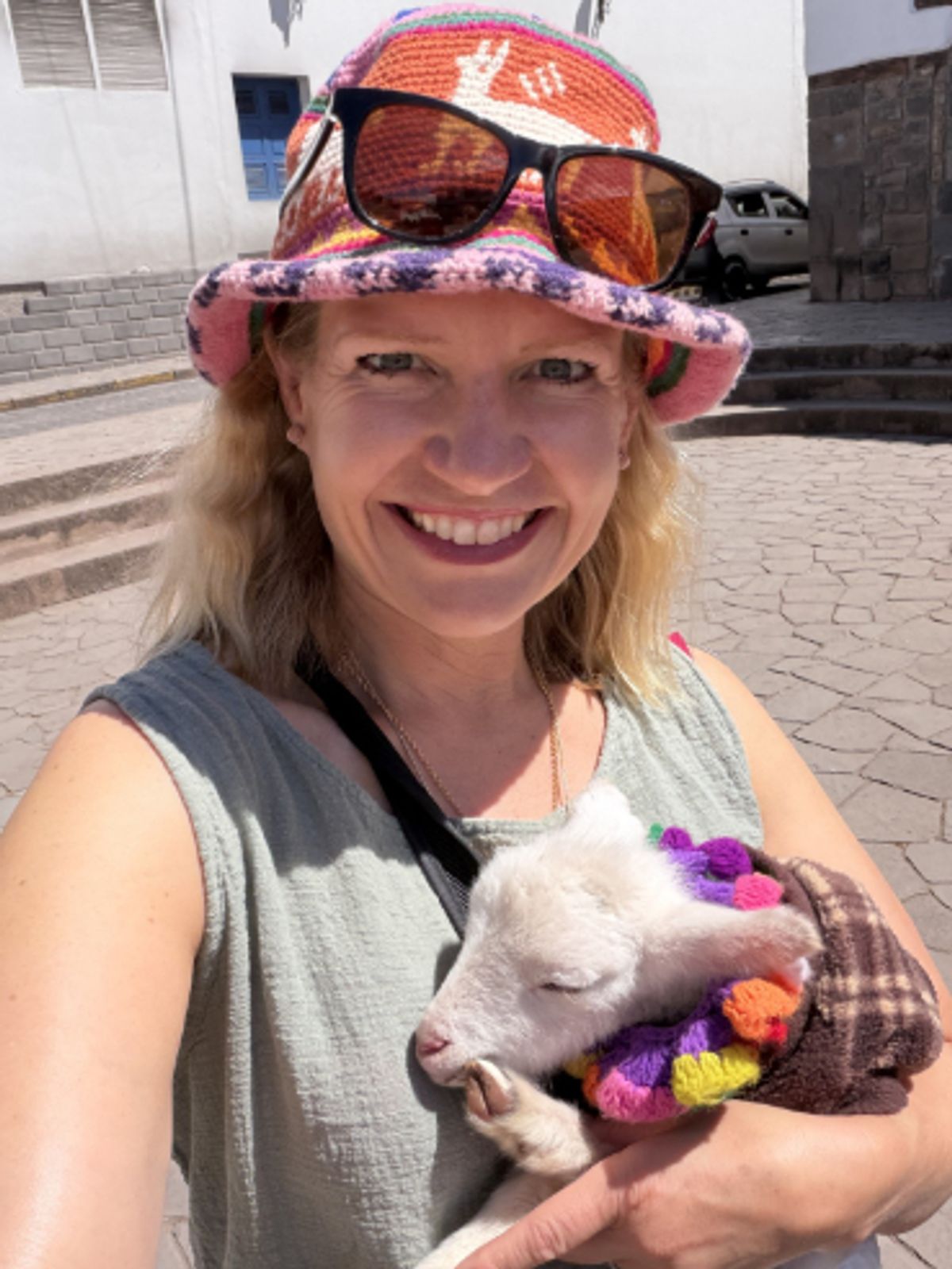 Smiling person outdoors holding a small white lamb, wearing a patterned bucket hat and large red-tinted sunglasses on a cobblestone street.