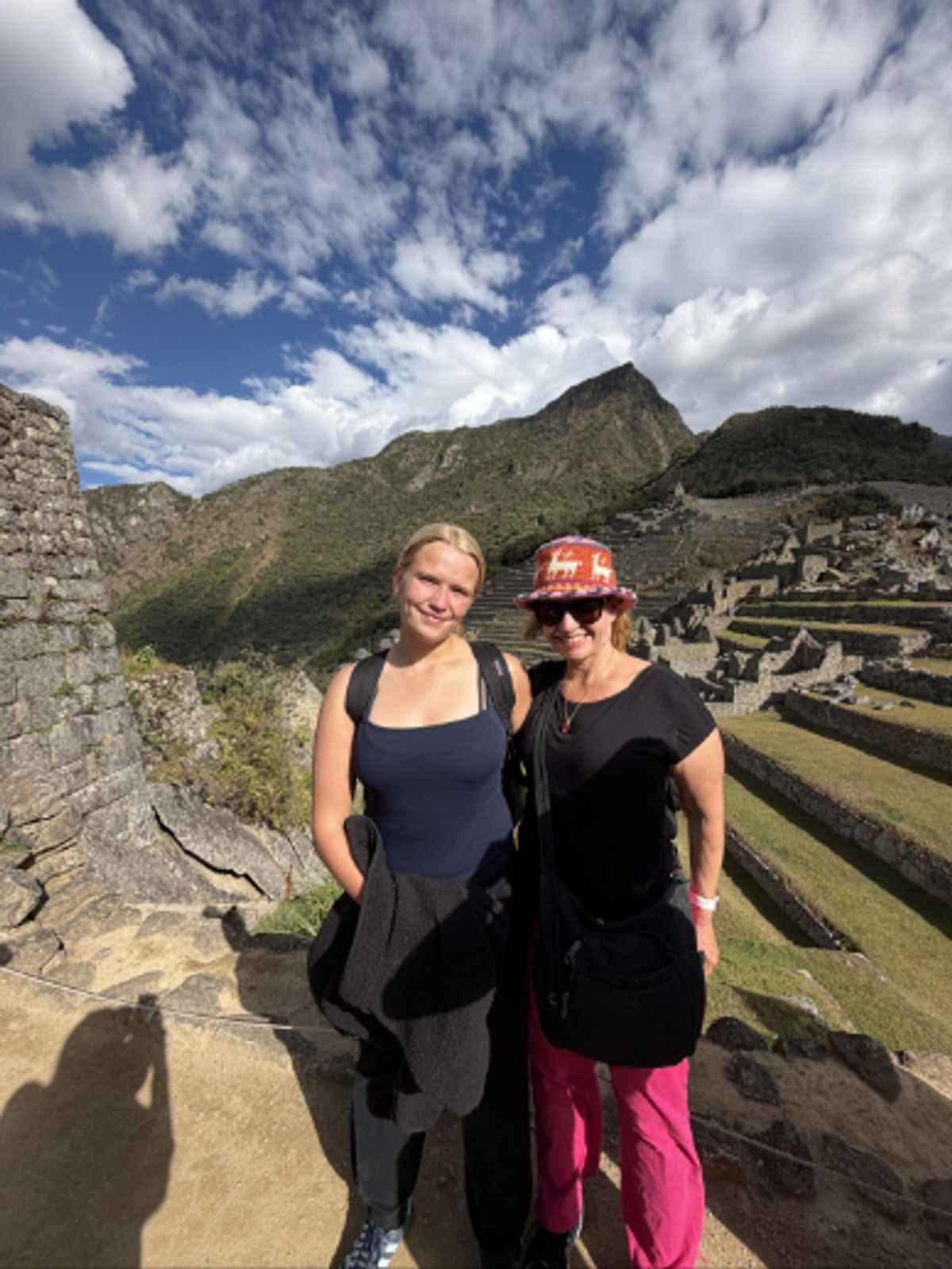 Two people posing on a stone walkway at a mountain archaeological site, with terraced ruins and green hills in the background under a partly cloudy blue sky.