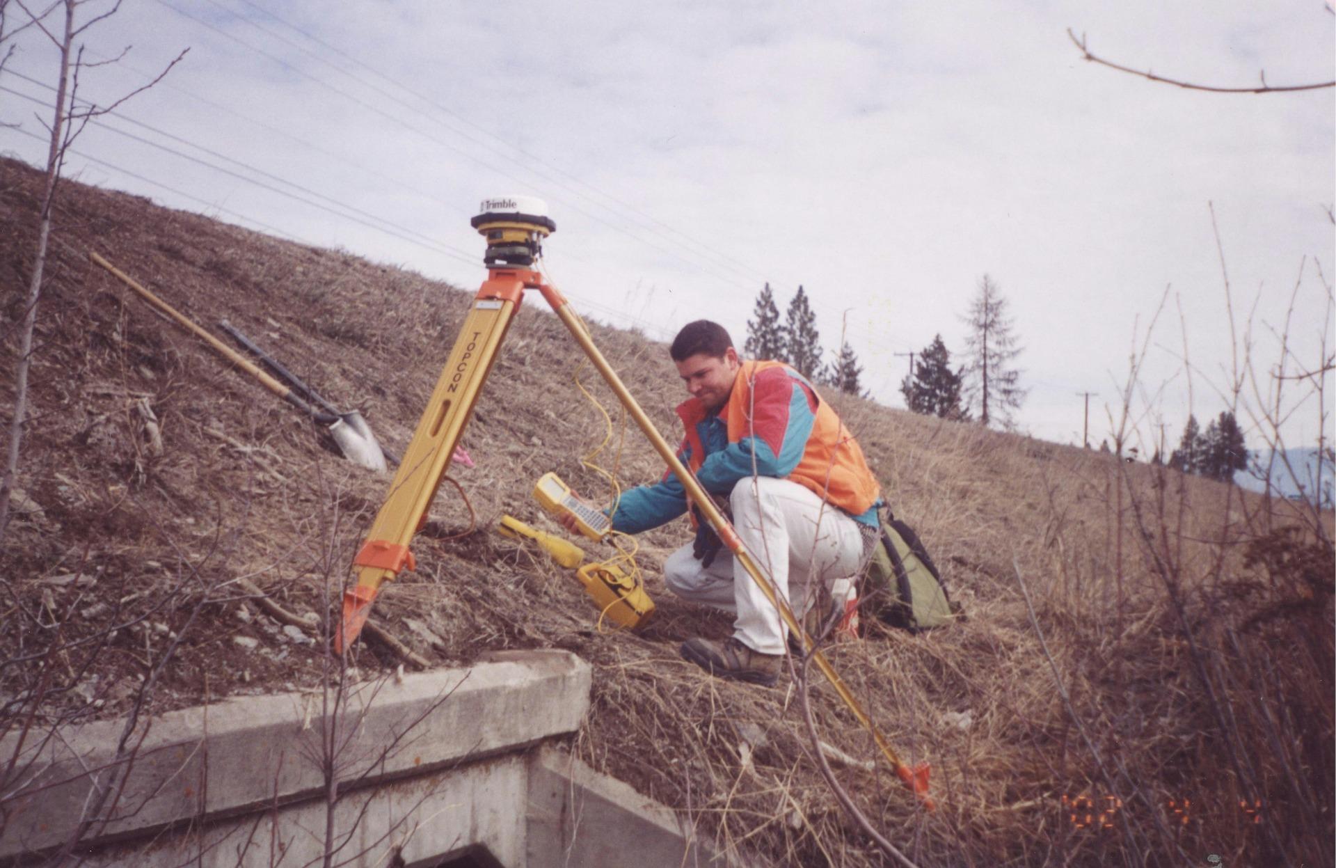 Sample photo: a surveyor setting up a GNSS receiver on a tripod beside a culvert