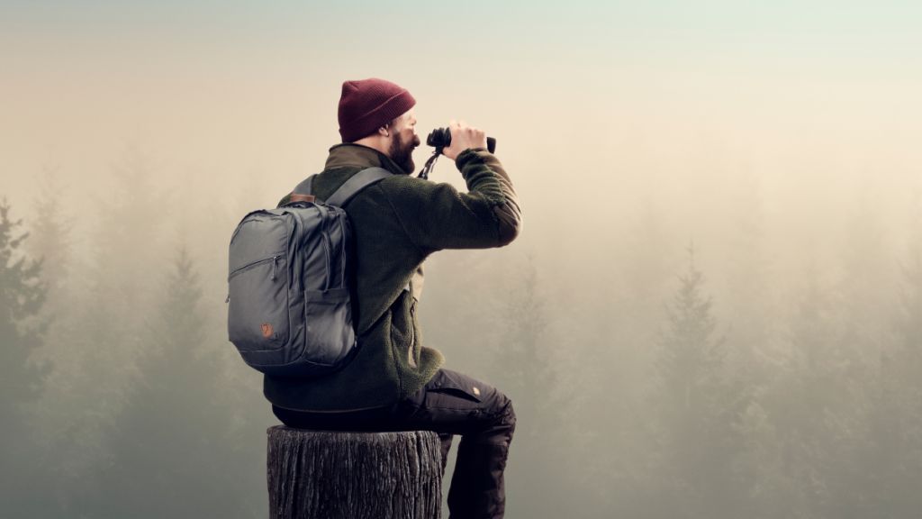 A person in a green jacket and red beanie sits on a log, using binoculars to observe a misty forest landscape.
