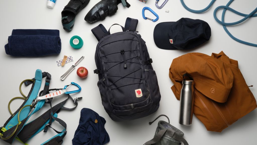 A flat lay of outdoor gear including a backpack, water bottle, towel, climbing harness, cap, and various accessories.