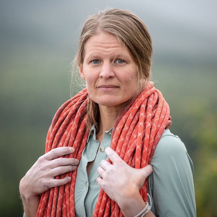A woman with long hair, wearing a light blue shirt and an orange scarf, stands outdoors with a blurred green background.