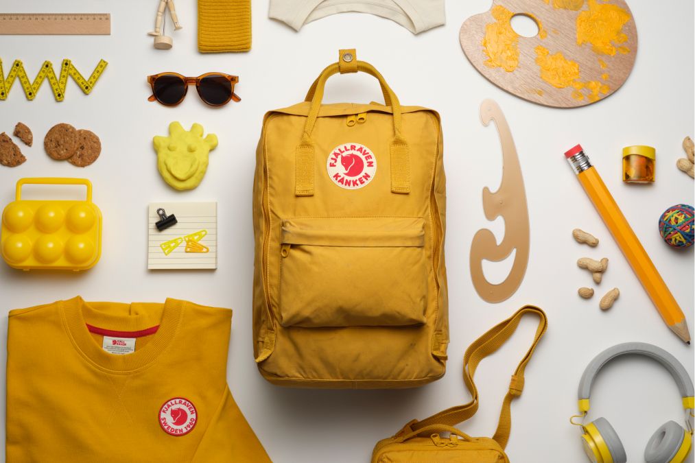 A flat lay of a yellow backpack surrounded by various colorful items, including cookies, a palette, sunglasses, and stationery.