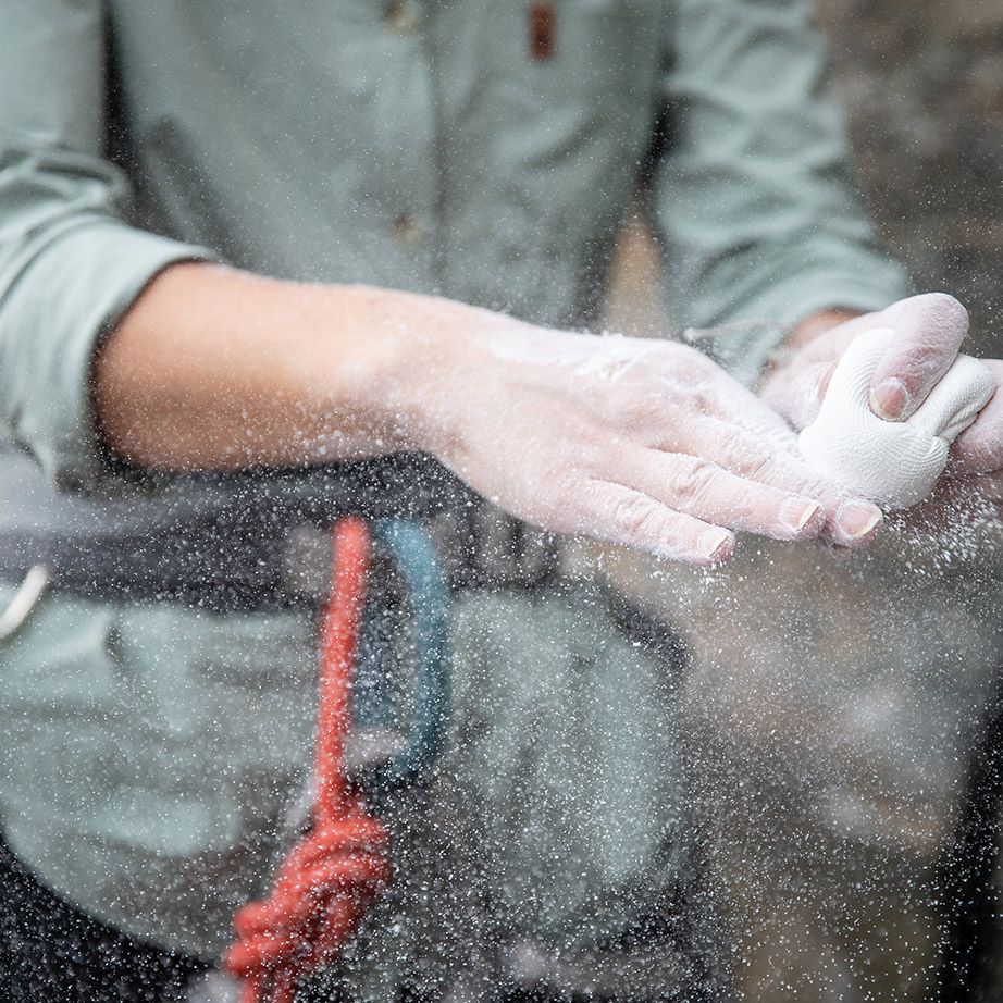 A climber applies chalk to their hands, creating a cloud of powder, with climbing gear visible in the background.