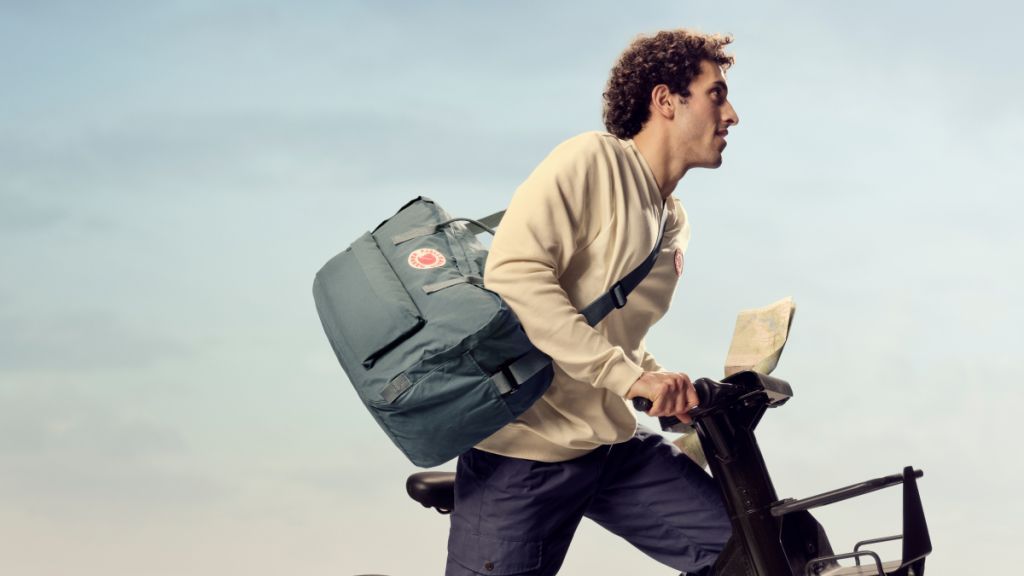 A man rides a bike, wearing a light sweater and a large backpack, holding a map against a cloudy sky backdrop.