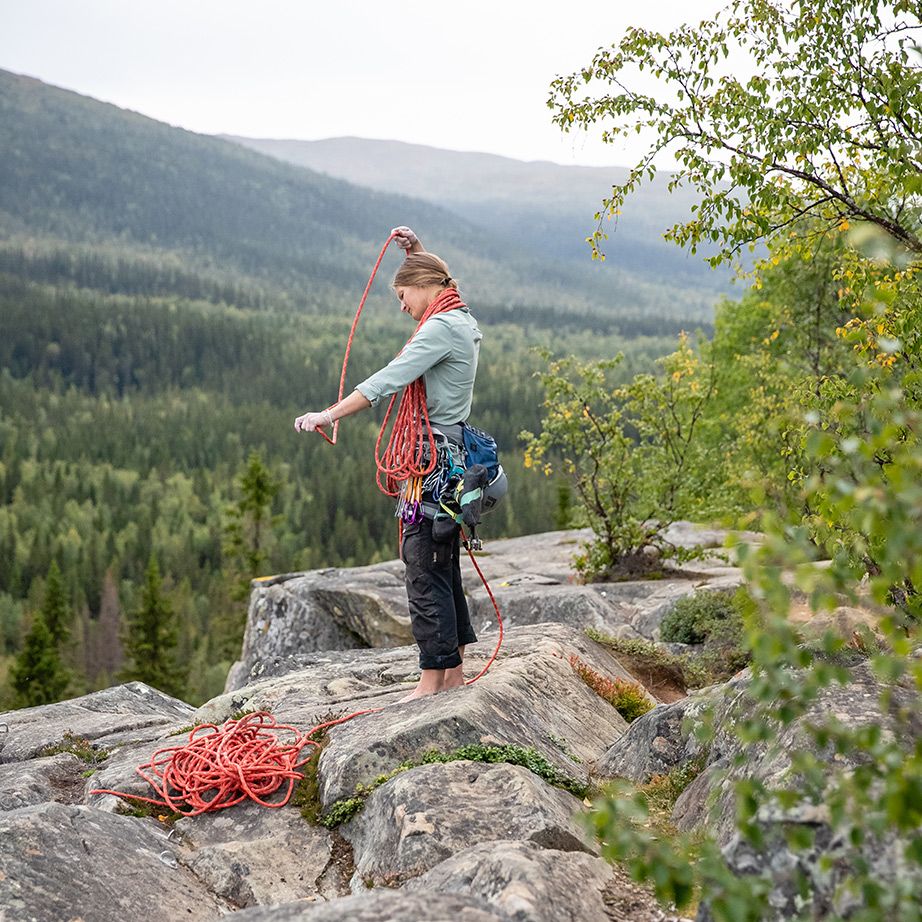 A climber stands on rocky terrain, preparing climbing ropes, with a lush forested valley in the background.