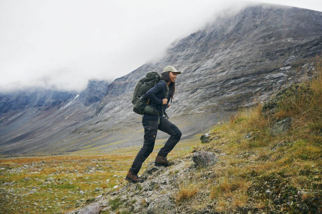 A hiker ascends a rocky slope in a mountainous landscape, wearing a backpack and outdoor gear under a cloudy sky.
