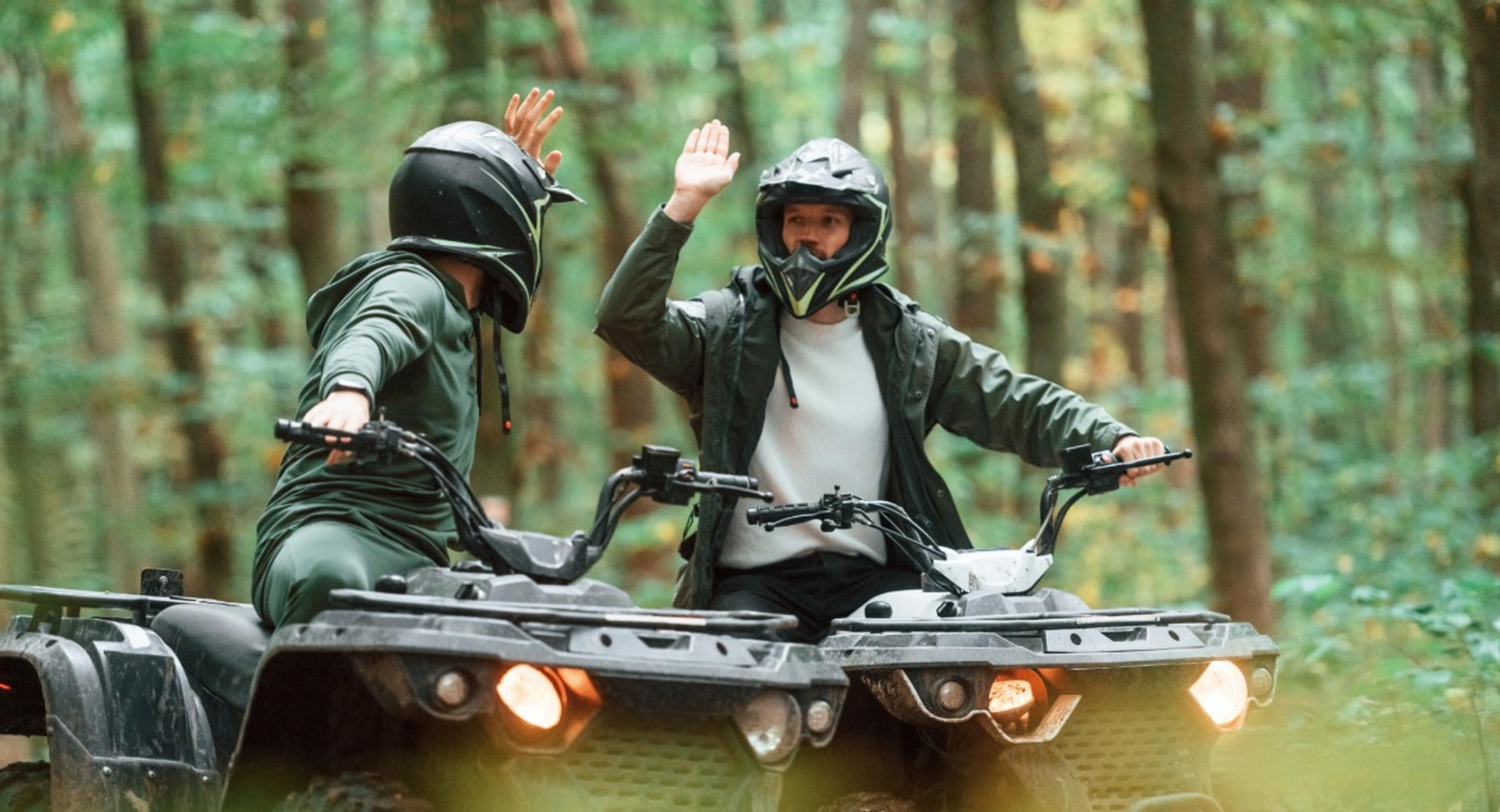Two ATV riders in helmets giving high-five on forest trail, demonstrating safe recreational ATV riding practices covered by liability insurance
