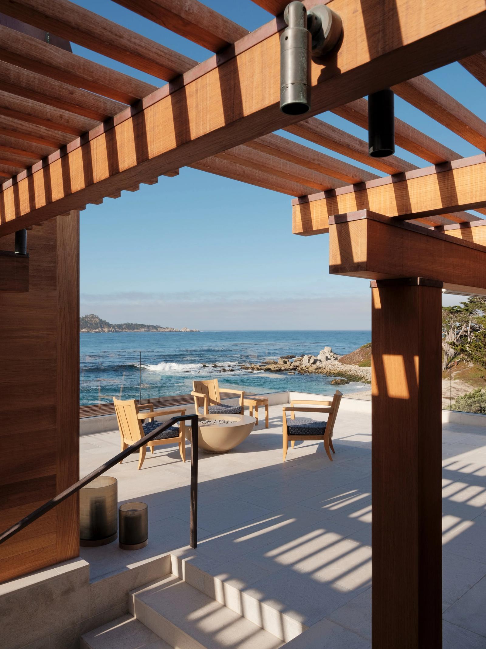 Coastal patio framed by a wood pergola, featuring outdoor lounge chairs arranged around a round concrete fire pit. Steps lead down to the seating area, which overlooks rocky shoreline and ocean views under clear skies.