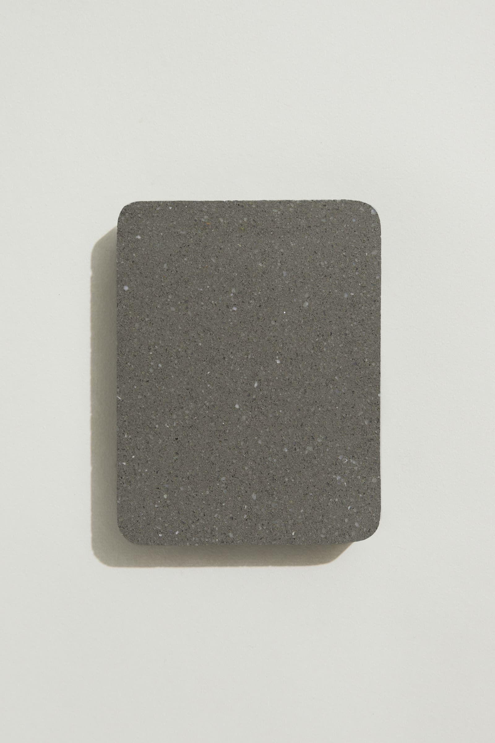 A rectangular sample of smooth dark grey concrete with grey, white and black terrazzo speckling, and rounded corners. Photographed on a white background with a cast shadow