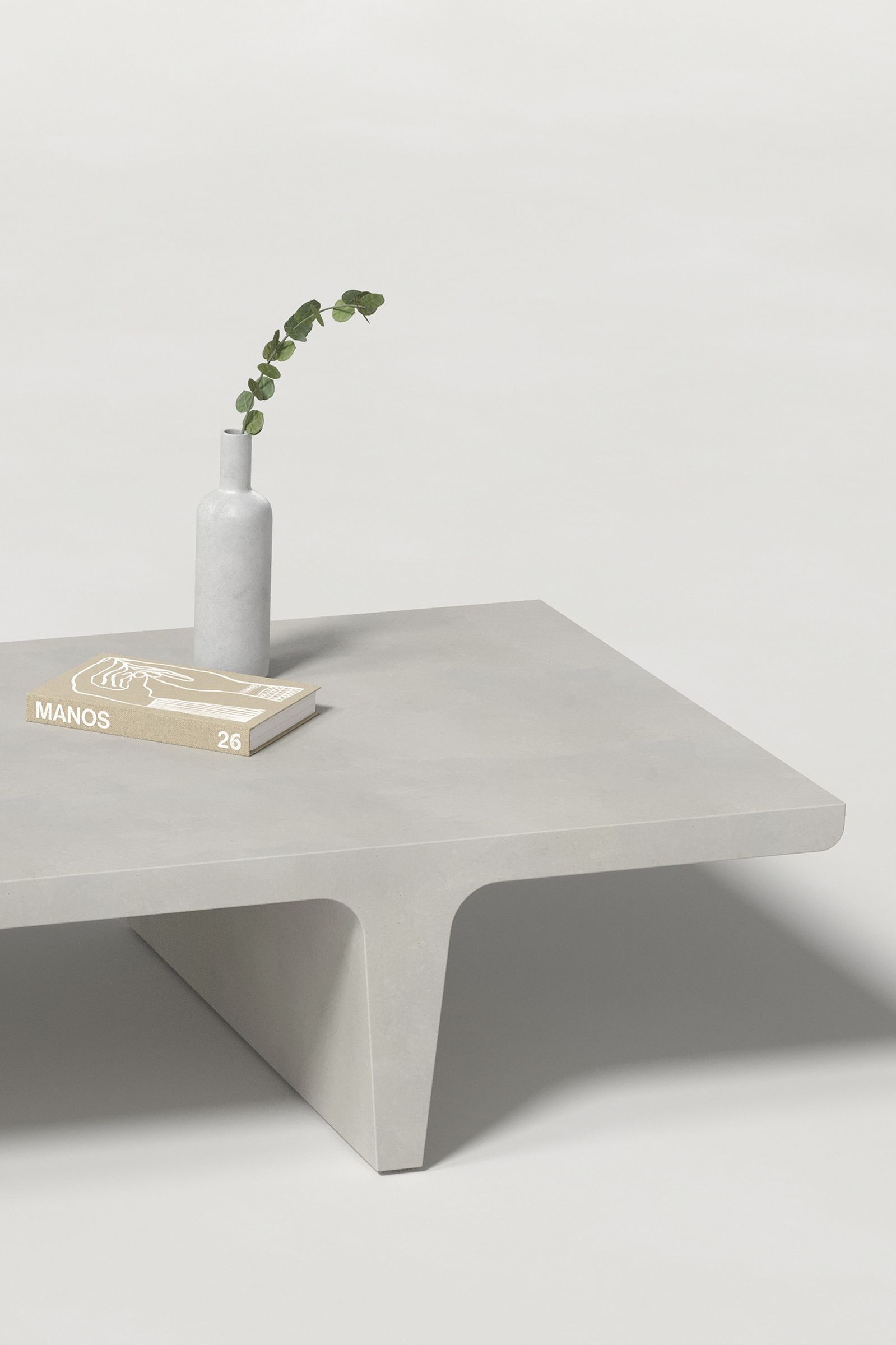 Close-up of a rectangular light grey concrete coffee table with sculptural double tee legs, styled with a neutral-toned book and a slender vase holding a eucalyptus, set against a clean white background.
