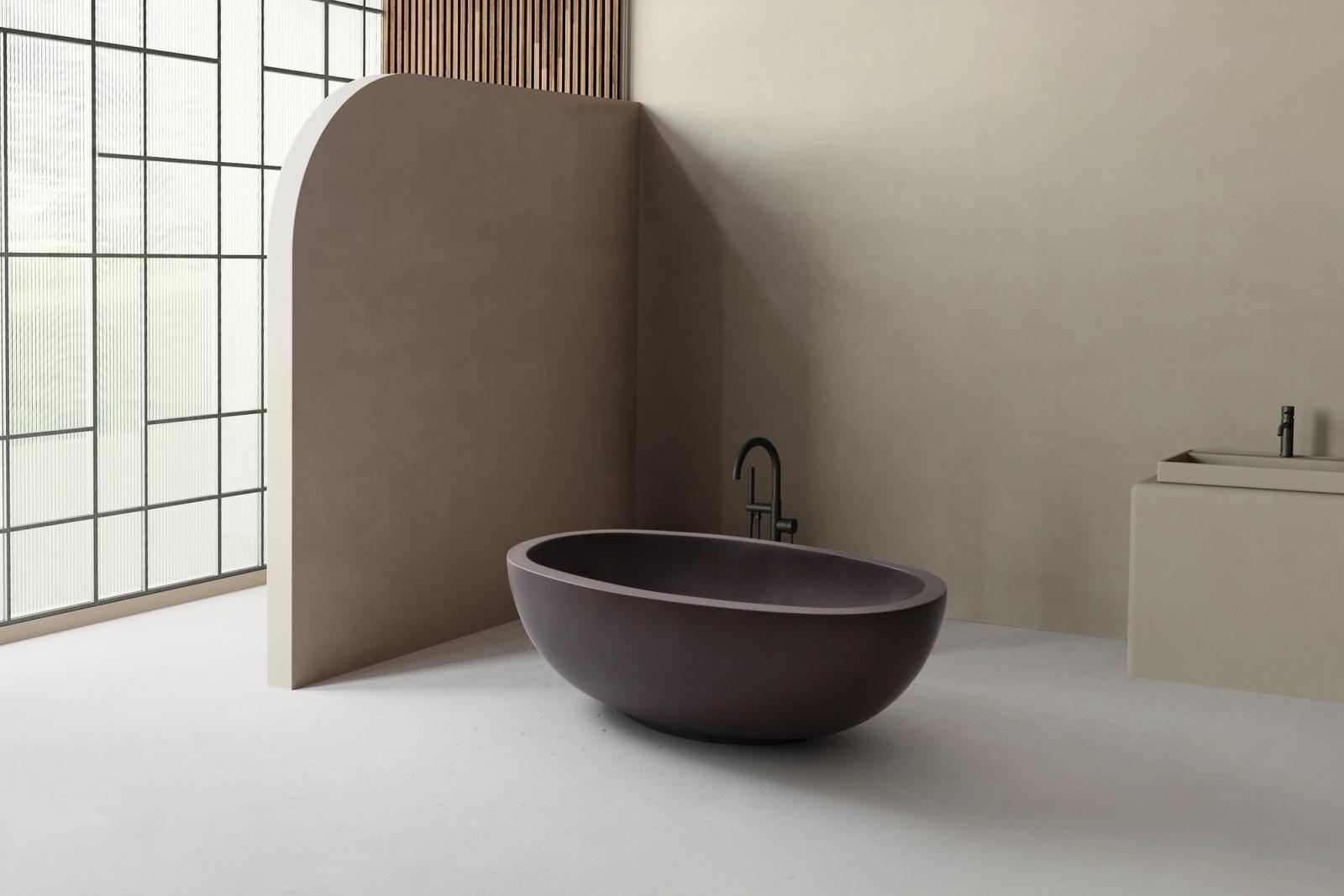 A modern bathroom with a deep, oval-shaped freestanding bathtub in a rich plum tone. The space features minimalist beige walls, a curved privacy partition, a large frosted glass window, and a coordinating sink with black fixtures.