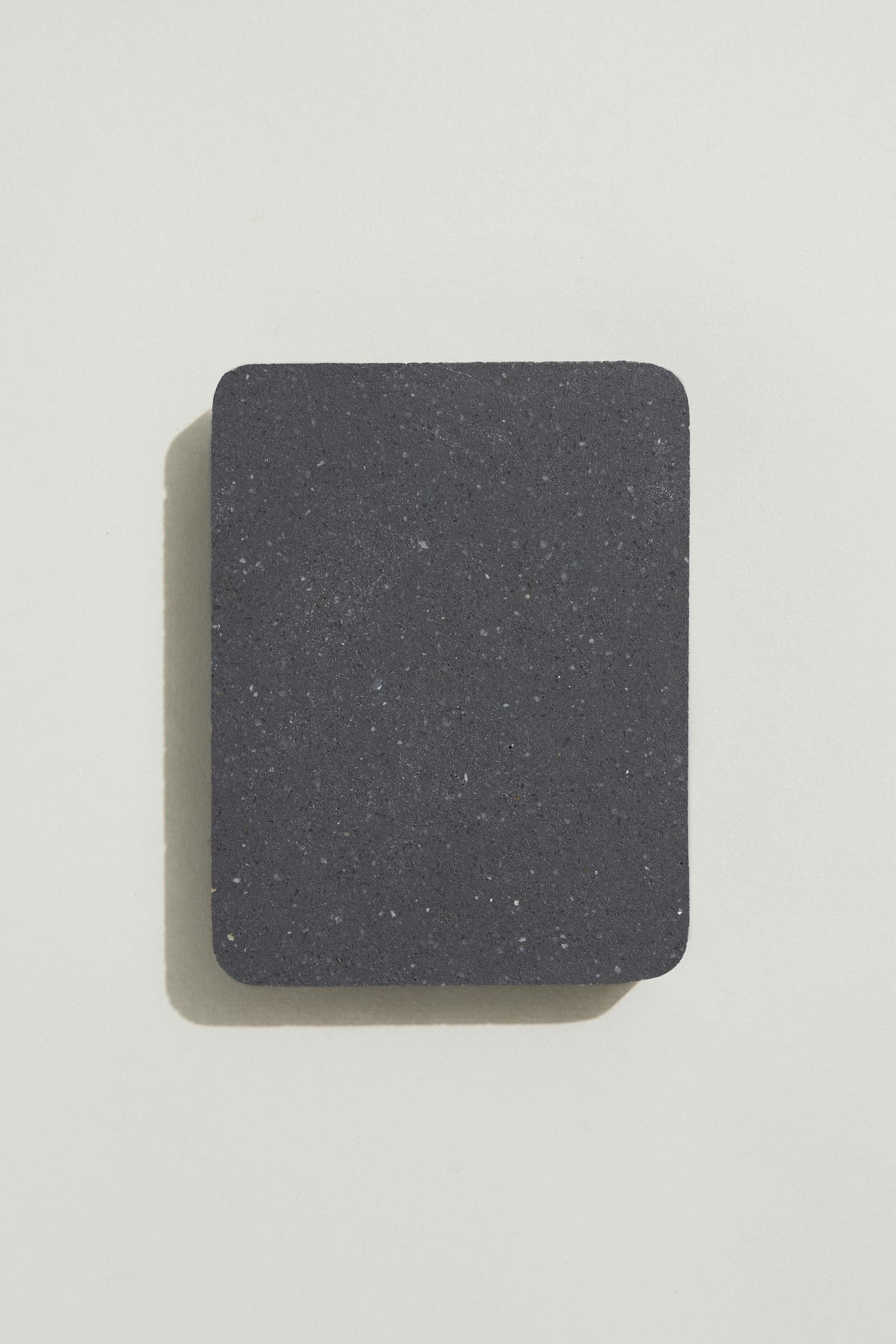 A rectangular sample of smooth charcoal concrete with grey, white and black terrazzo speckling, and rounded corners. Photographed on a white background with a cast shadow