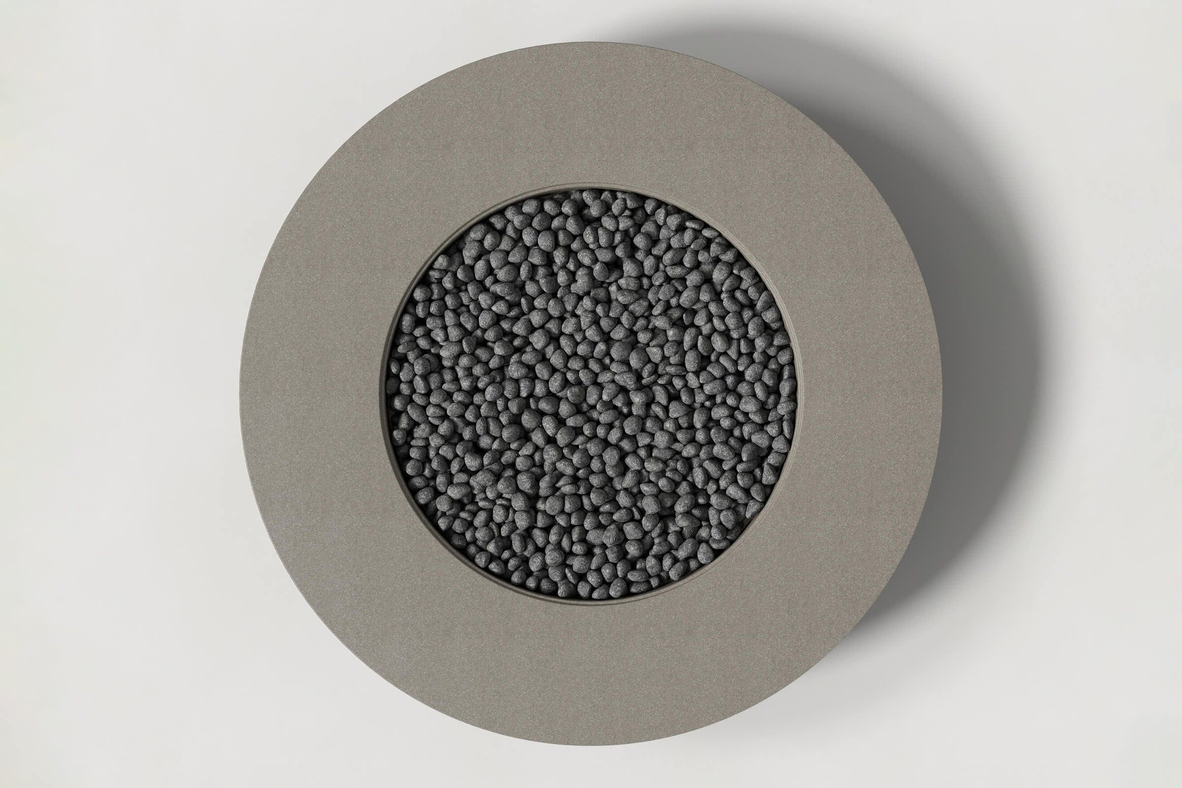 Top-down view of a circular dark grey concrete fire pit with thick walls and a circular well filled with volcanic rocks. Shown against a white background with slight shadows.