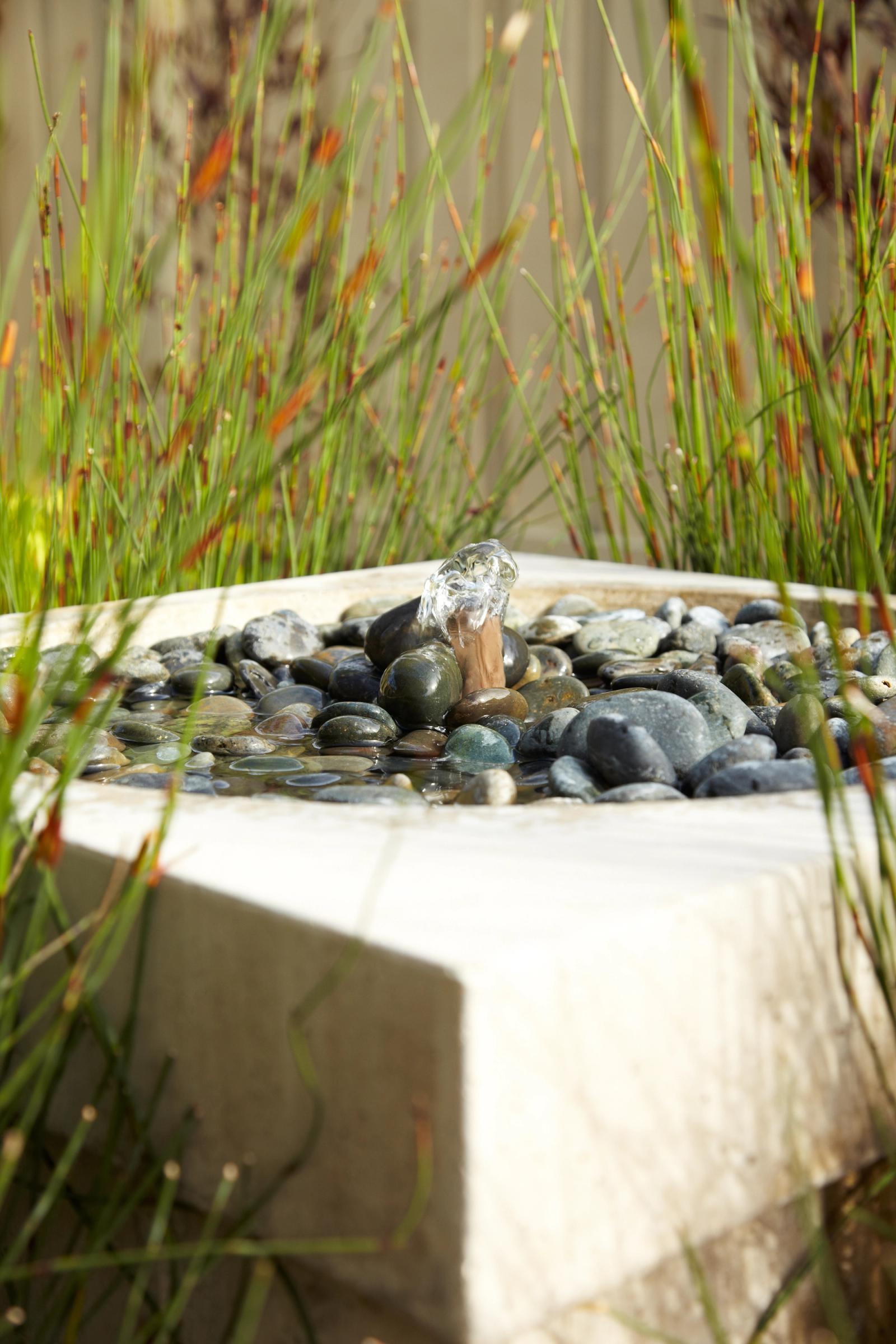 Square concrete water basin filled with smooth river stones, surrounded by tall grasses in a landscaped garden setting.