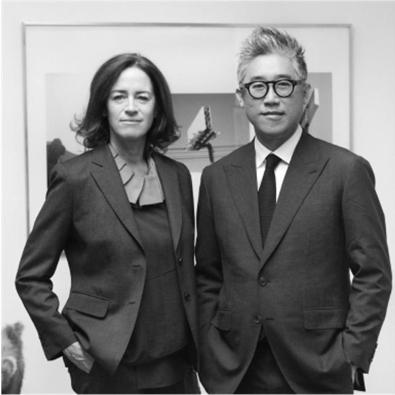 A black and white portrait of two people standing against a light wall. Both wear formal suits and project a professional demeanor. Artwork is partially visible behind them.